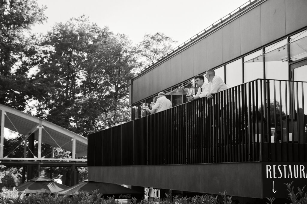 Zdjęcie przedstawia grupę mężczyzn w restauracji na tarasie z widokiem na drzewa, w eleganckich strojach, podczas rozmowy.