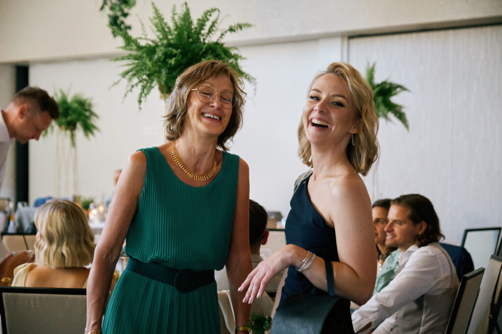 Dwie kobiety ucieszone, świętujące na weselu, w eleganckich sukienkach, w otoczeniu gości.