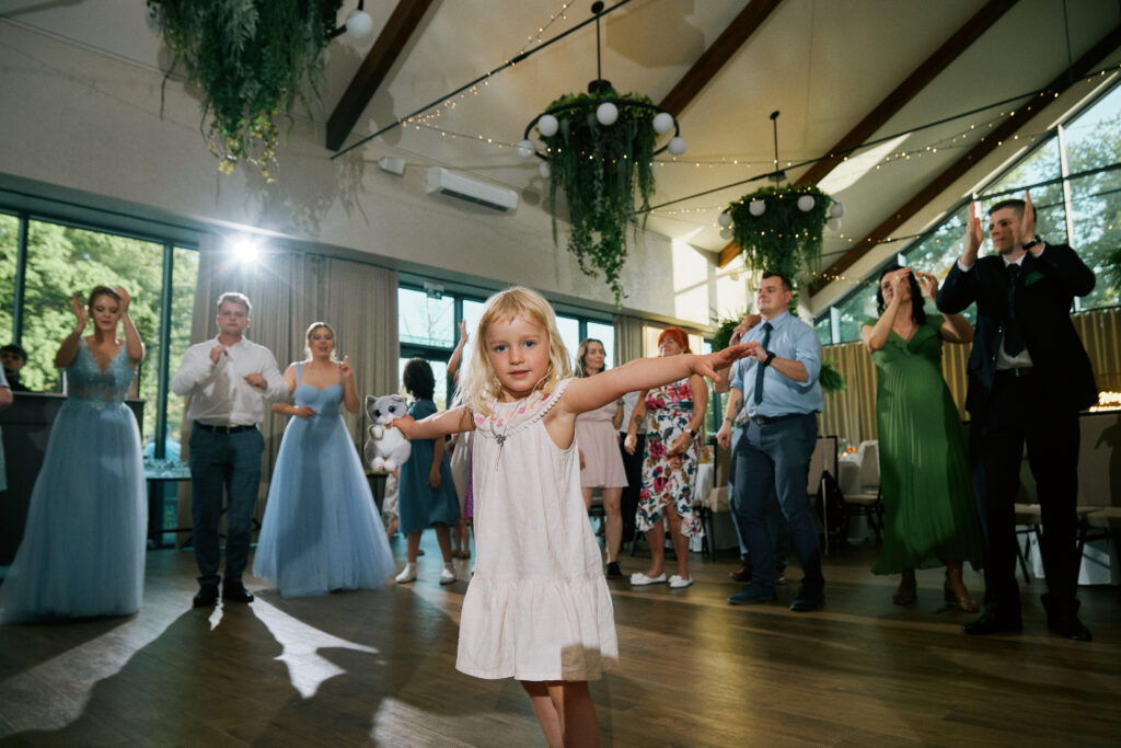 Piękna dziewczynka tańczy na weselu, w eleganckim salonie, dekoracje i ludzie w tle.