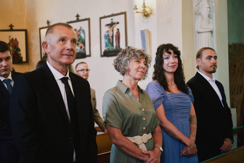 Alt: Grupa ludzi na ceremonii religijnej w kościele, skupieni, elegancko ubrani, z obrazami religijnymi na ścianie.