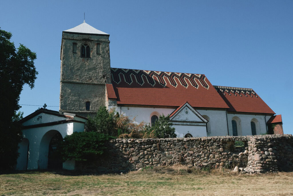 Stary kościół z czerwonym dachem i wieżą, otoczony kamiennym murem, na tle błękitnego nieba.