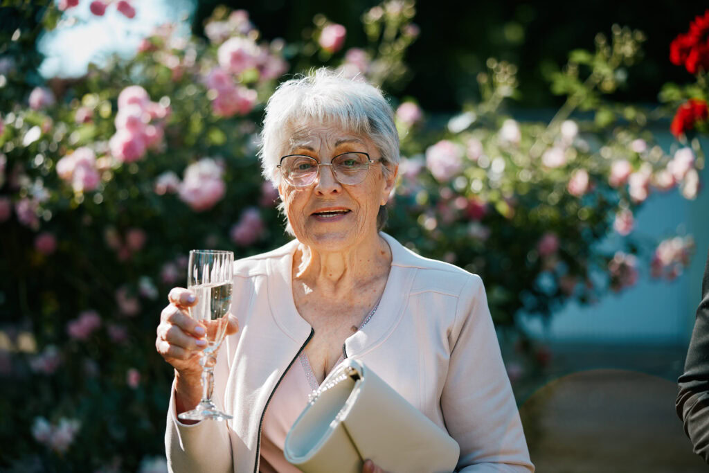 Przedstawiająca starszą kobietę z kieliszkiem szampana, na tle kwitnących różowych krzewów i zieleni, z uśmiechem na twarzy.