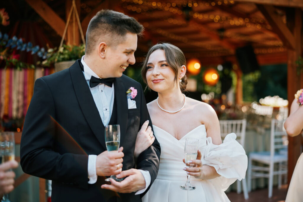 Alt="Para młoda podczas wesela, uśmiecha się do siebie przy lampkach szampana".