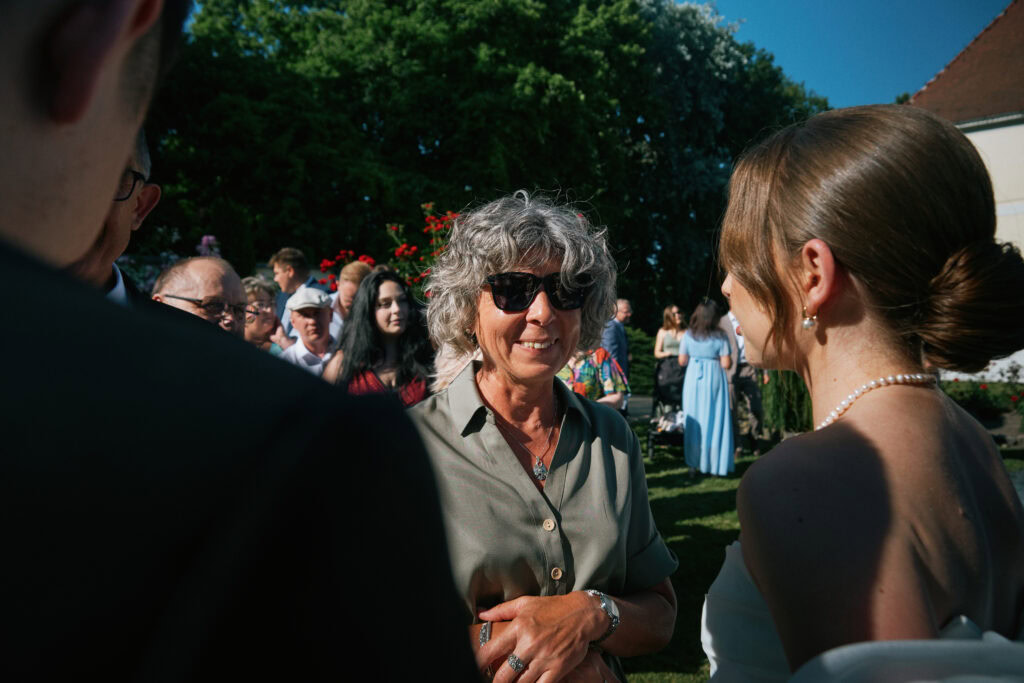 Górna część zdjęcia ukazuje gości na weselu w plenerze, skupieni na rozmowach, z pięknymi dekoracjami i zielenią w tle.