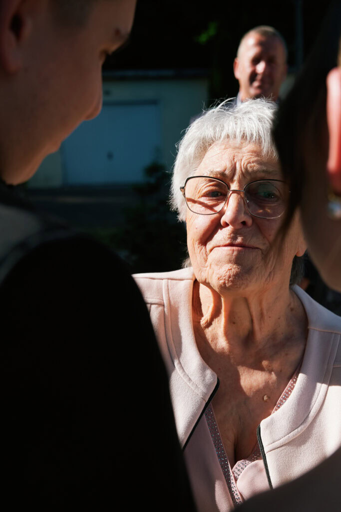 Uśmiechnięta, starsza kobieta w okularach, otoczona bliskimi podczas rodzinnej uroczystości.